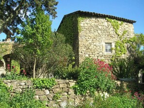 Surrounded by lush greenery and flowering plants, this historic property features a stone facade and a tiled roof.