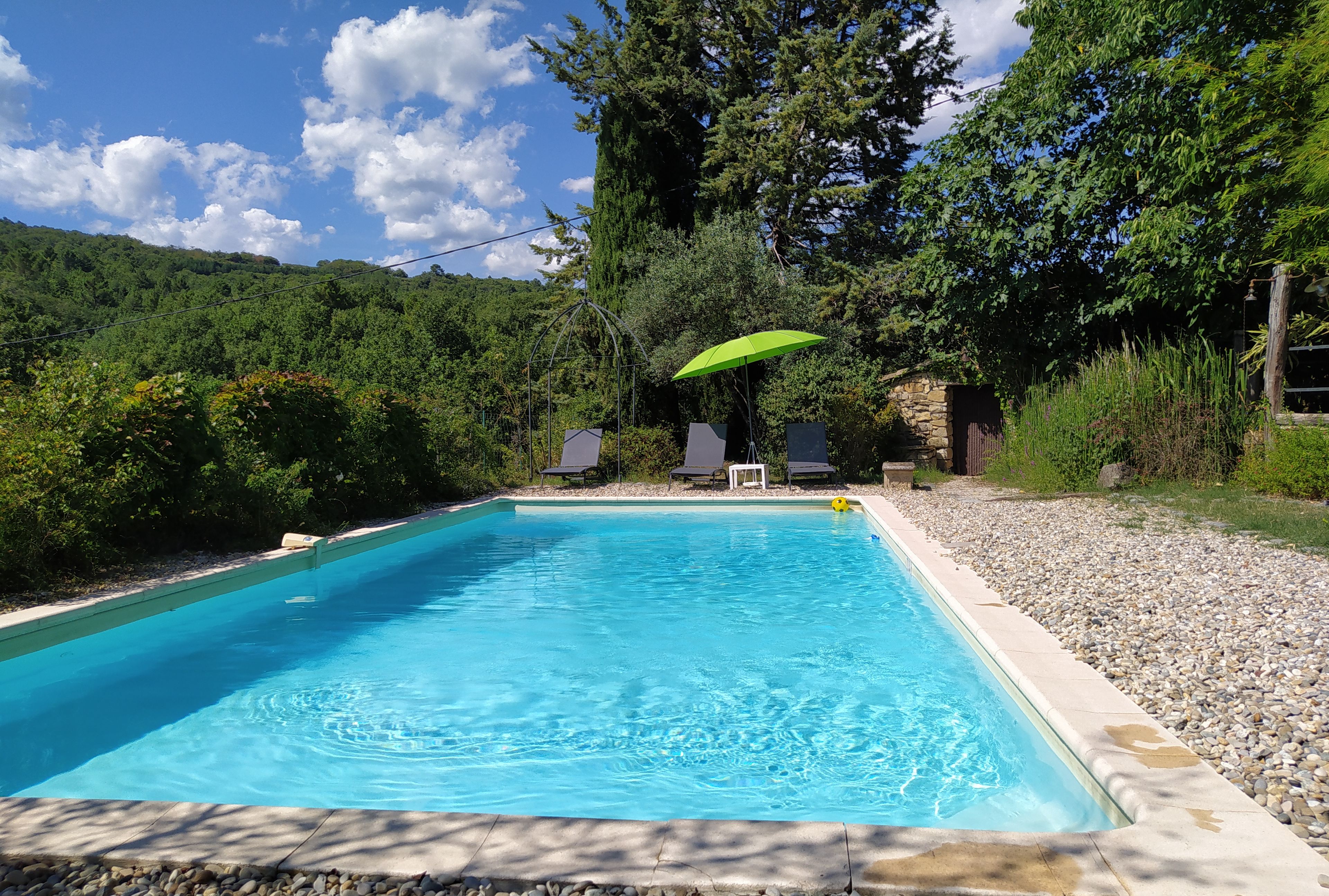 A rectangular swimming pool with clear blue water, surrounded by a pebble border. There are two lounge chairs and a green umbrella under a pergola. The pool is set in a landscaped area with trees and shrubs.