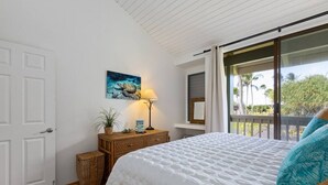 Un dormitorio con una cama grande, una mesita de noche con lámpara, un cuadro y vistas a palmeras a través de un ventanal corredero.