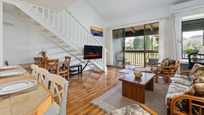 Un salón amplio con suelo de madera, zona de comedor con una mesa dispuesta para dos personas, sofá y televisor montado en la pared.