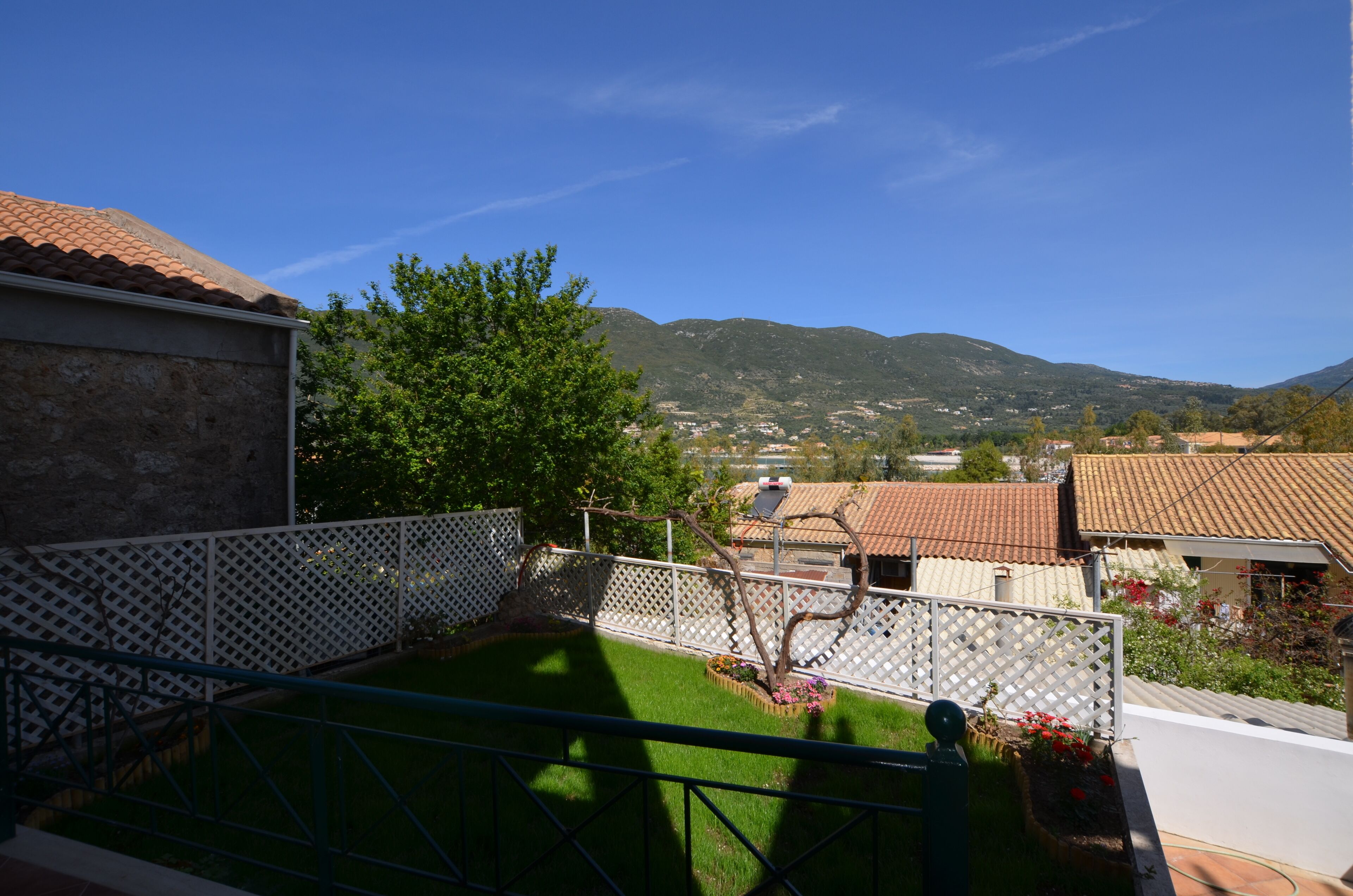 A residential area with a view of mountains, a fenced yard with a tree, and houses with red-tiled roofs.