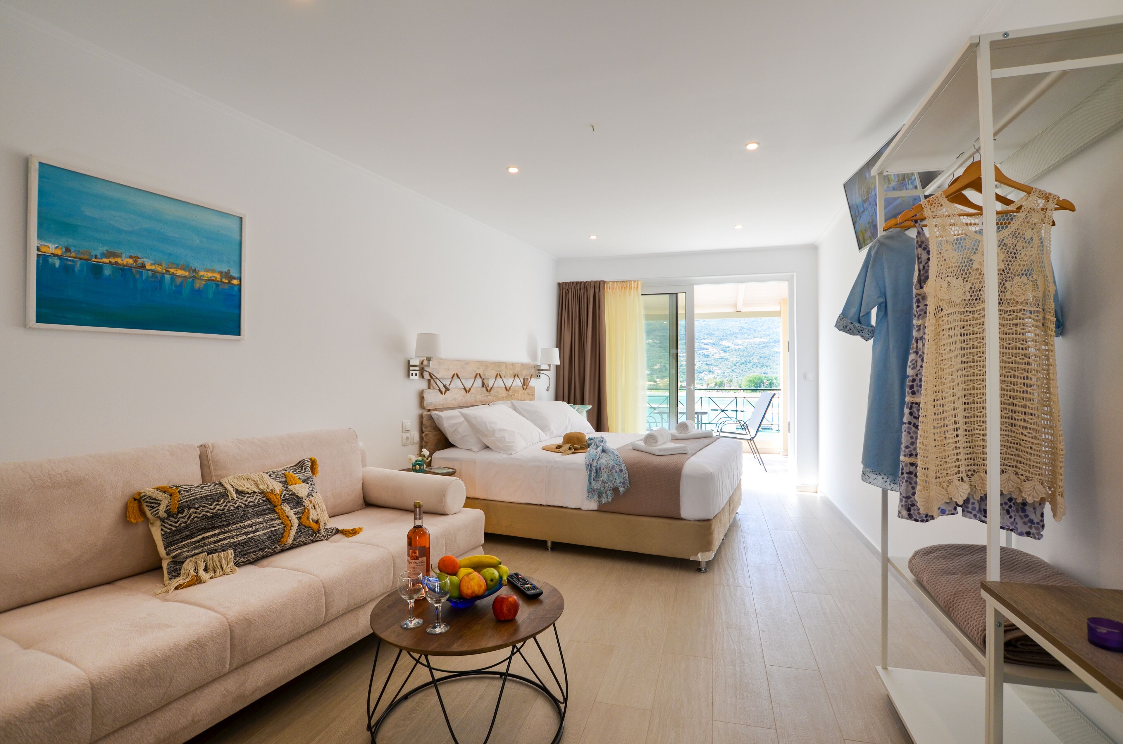 Une chambre d’hôtel avec un lit, un canapé, une table basse avec des fruits et une bouteille, et un balcon où sont suspendus des vêtements.