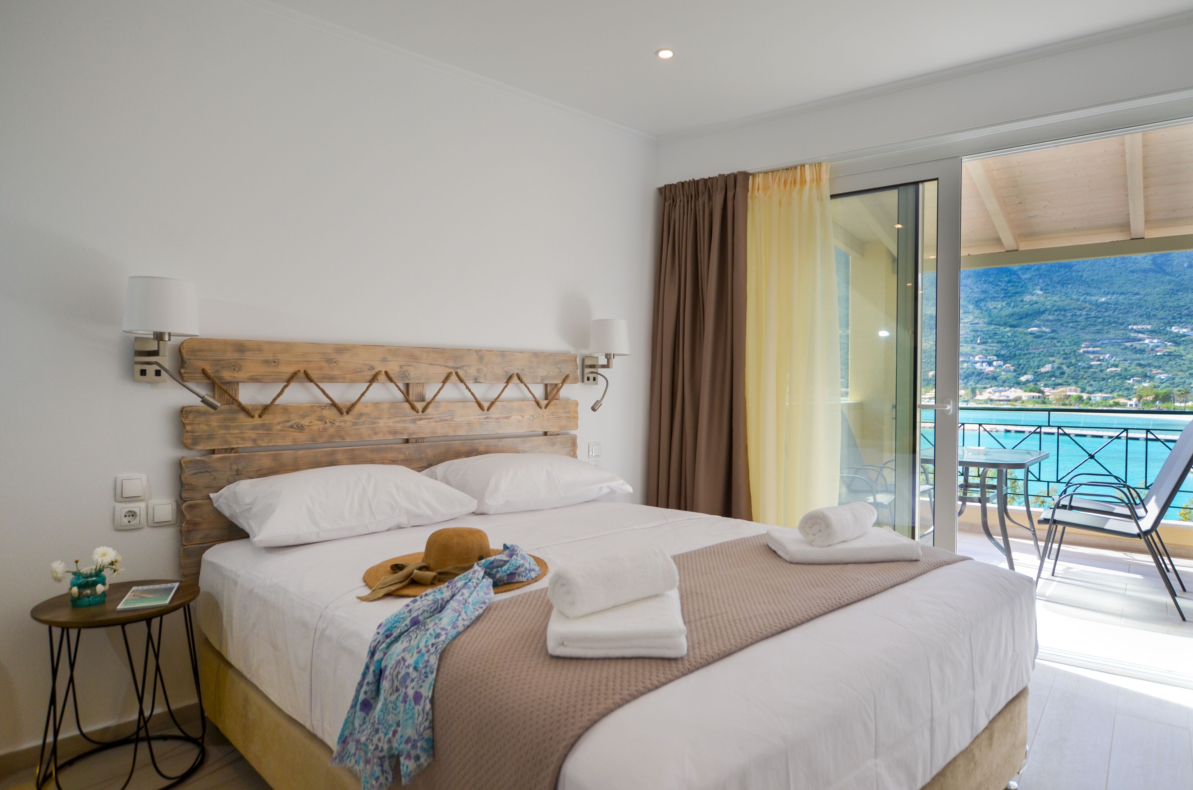 Habitación de hotel con una cama grande, cabecera de madera, una mesita con un sombrero y flores, y un balcón con vistas al mar.