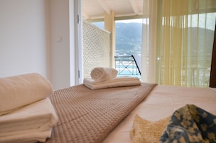 Habitación de hotel con cama, toalla doblada, manta con textura y vista al mar desde un balcón con barandilla.