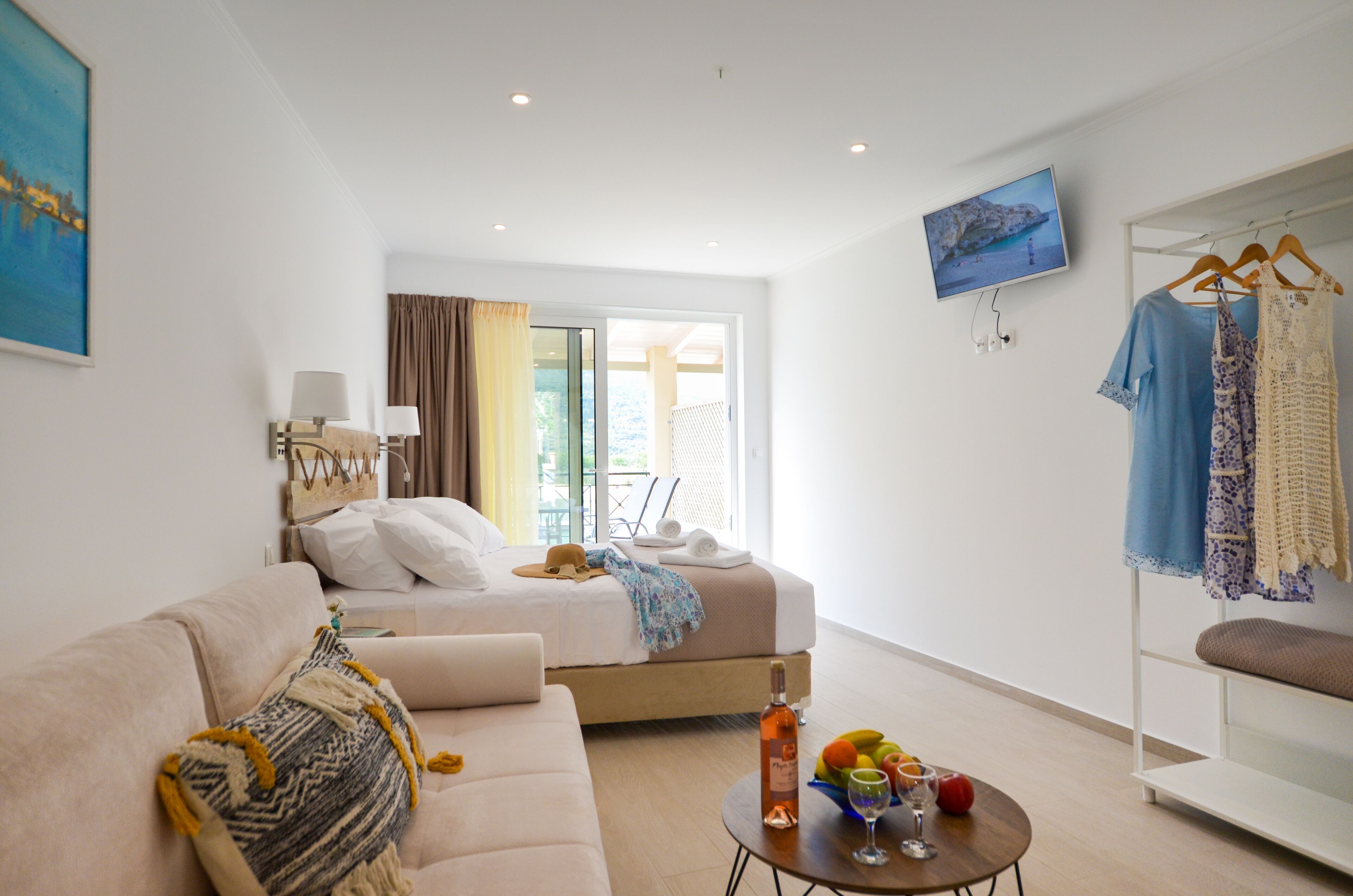 A modern hotel room with a bed, a sofa, a table with fruit and a bottle, and a clothing rack.