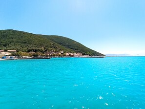 Un pueblo costero con edificios a lo largo del paseo marítimo, un mar azul cristalino y una colina con vegetación.