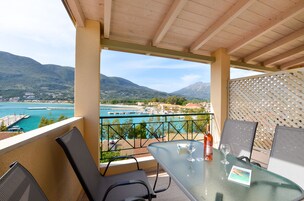Un balcón con una mesa de vidrio, dos sillas y una botella de vino. Se aprecia una vista de la costa con montañas y un puerto deportivo.