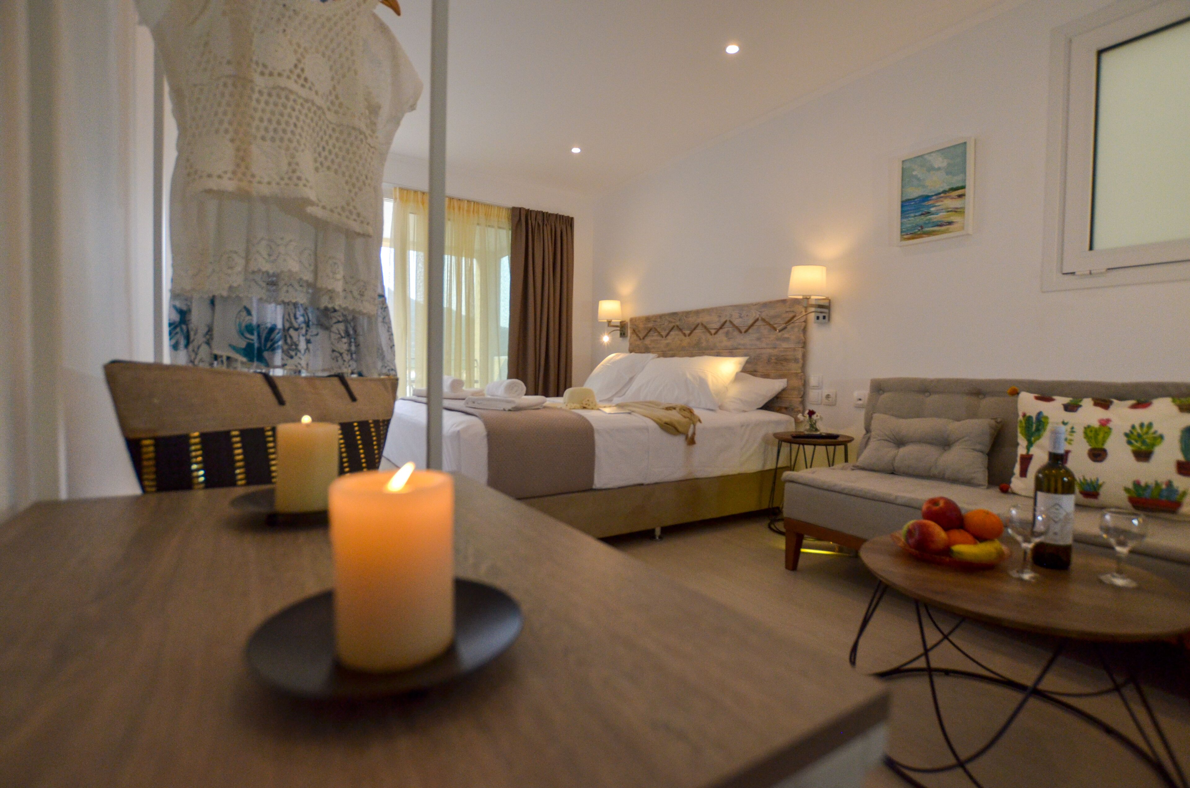 Habitación de hotel con cama, sofá, mesa de comedor con vela, y una mesita con frutas y copas de vino.