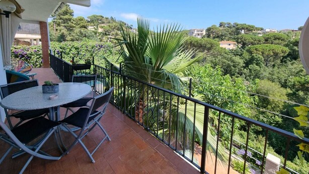 A balcony with a round table and chairs, surrounded by a metal railing, overlooking a lush green landscape with trees and buildings in the distance.