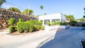 A modern building with a stone retaining wall, surrounded by well-maintained shrubs and a clear sky. - 3082 RES LOMAS DE CAMPOAMOR (Orihuela Costa)