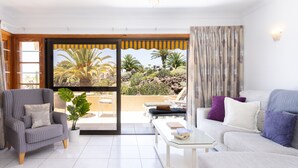 Una sala de estar con sofá, sillón y una mesa de centro. Se aprecia una ventana grande con vistas a palmeras y un balcón.