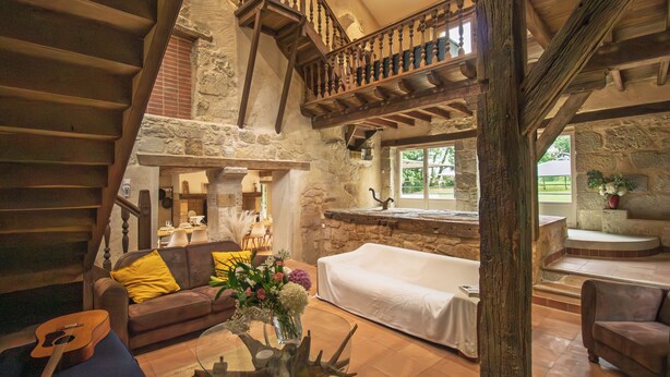 Un intérieur rustique avec un escalier en pierre, des poutres en bois, un canapé blanc et une table basse en verre ornée de fleurs.