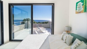 A bedroom with a large bed, a view of the sea, and a balcony with lounge chairs.