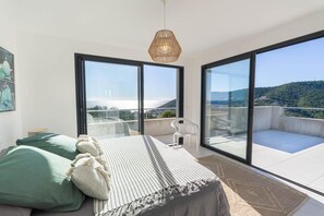 Camera da letto con un letto, cuscini, comodino, lampada a sospensione e ampie porte scorrevoli in vetro che danno su un balcone con vista sulla città e sulle colline.