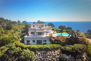 A modern two-story house with a pool, surrounded by lush greenery and overlooking the sea.