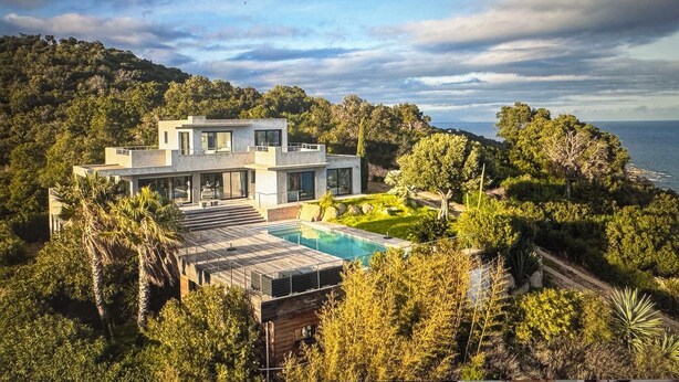A modern house with a pool, surrounded by lush greenery and overlooking a body of water.