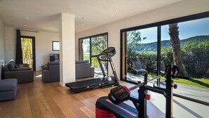 A spacious living area with a treadmill, a weight bench, and a large window offering a view of greenery.