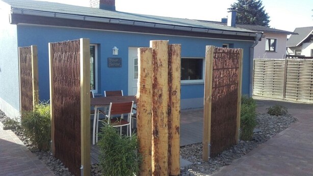 A blue building with a wooden fence, a patio with a table and chairs, and a brick pathway.