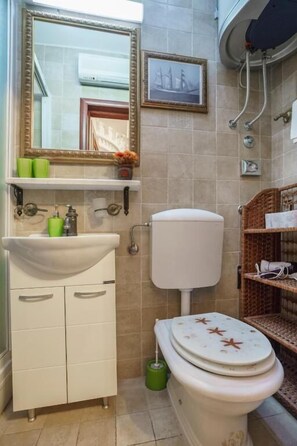A bathroom with a white sink, a toilet, and a shower. There are two framed pictures on the wall and a small shelf with green cups.