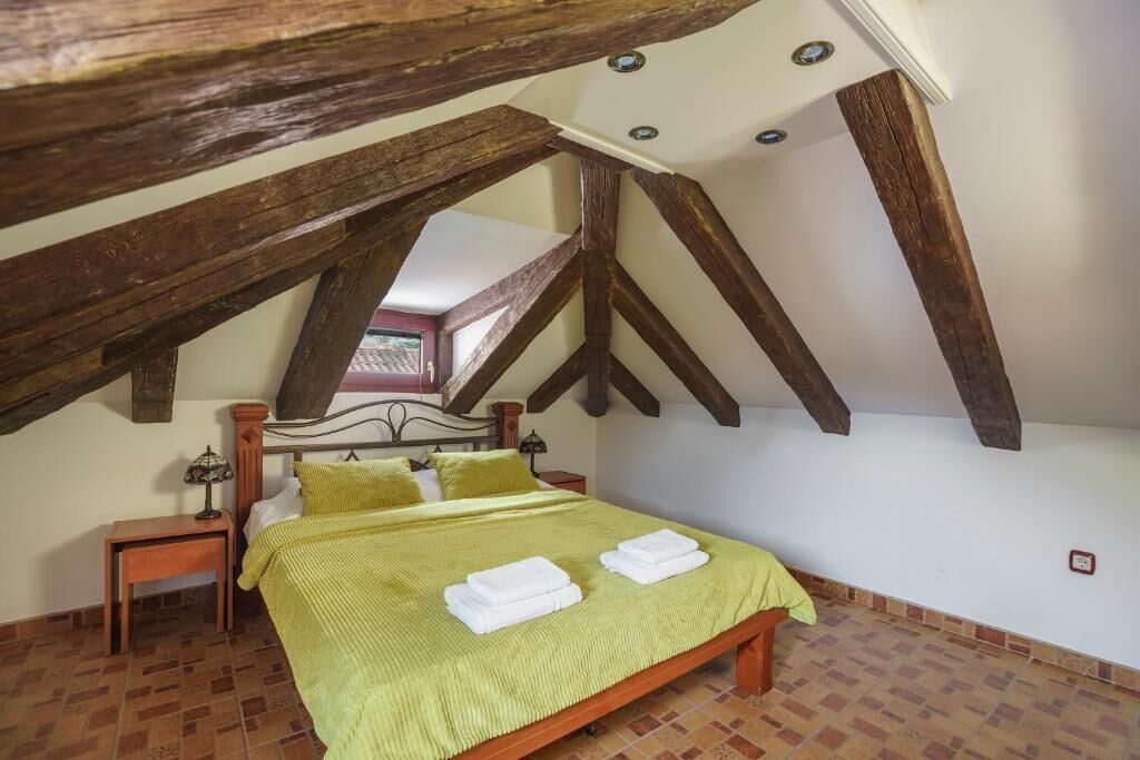 Ein Schlafzimmer mit schräg verlaufender Decke, Holzbalken, einem Bett mit grüner Bettwäsche und einem Nachttisch mit Lampe.