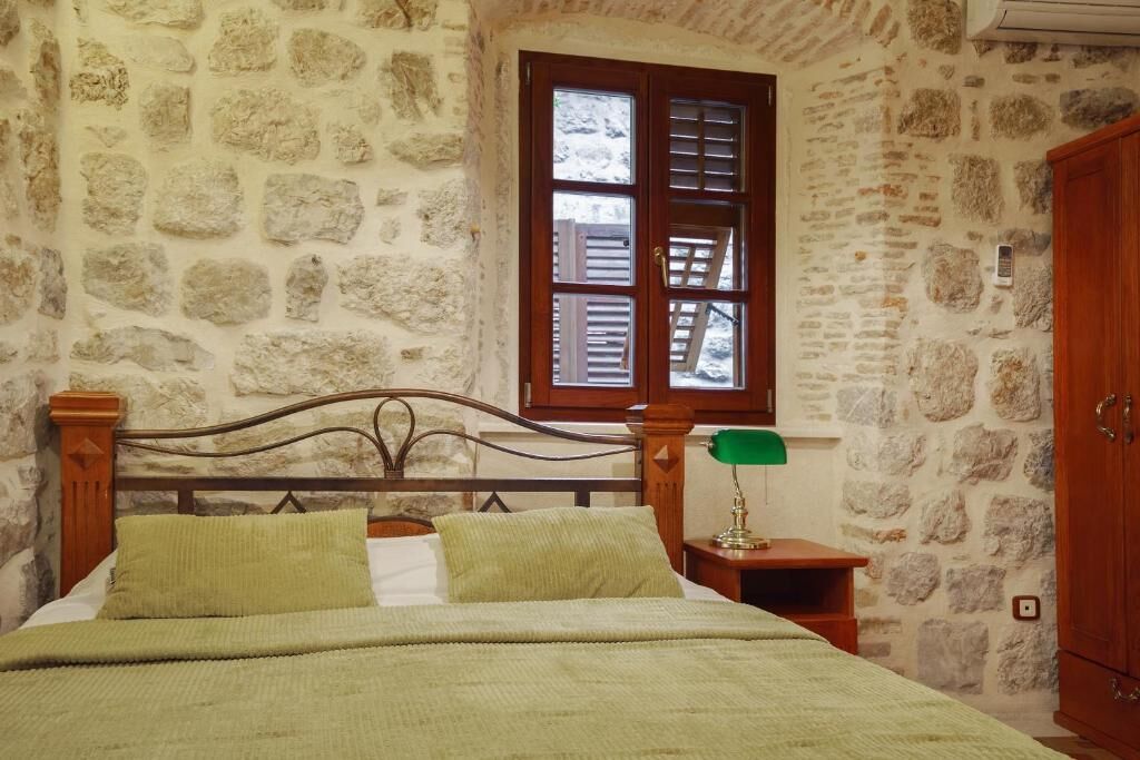 A bedroom with a stone wall, a wooden bed with green bedding, a nightstand with a lamp, and a window with shutters.