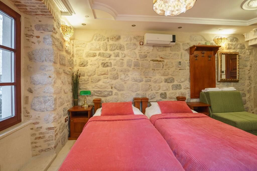 Une pièce aux murs de pierre, avec deux lits dotés de draps roses, une armoire en bois et une chaise verte.