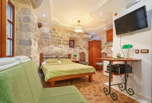 A room with a stone wall, a bed with green bedding, a TV, a desk with a lamp, and a chair.
