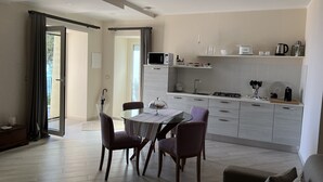 Une cuisine moderne avec des meubles blancs, un coin repas doté d’une table en verre et une vue sur l’extérieur grâce à la porte ouverte.