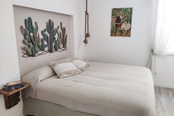 Een slaapkamer met een bed, een cactusschilderij, een schilderij en een klein plankje met boeken.