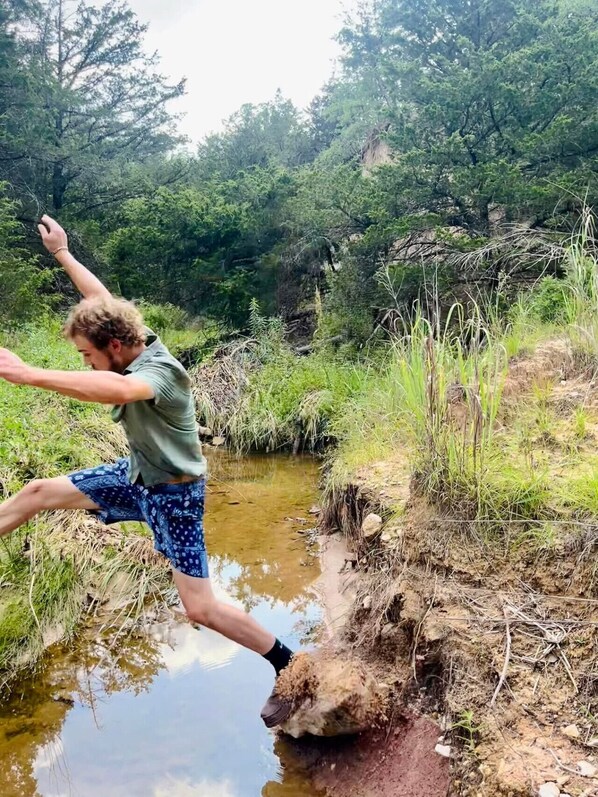 A person jumping over a small stream in a natural setting with trees and vegetation around. - Escape Velocity Dome (Saint Jo)