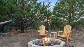 A fire pit with two wooden chairs and a small table, surrounded by trees. - Hippy Chick Yurt Cabin (Saint Jo)