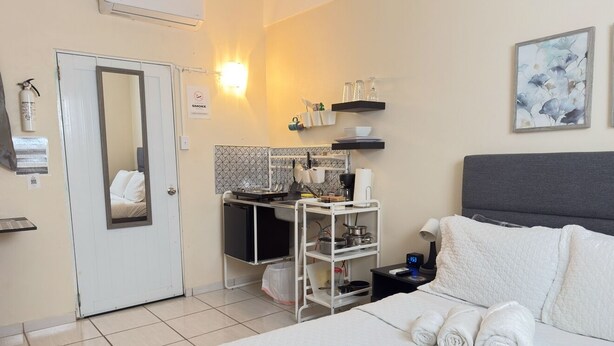 Quarto de hotel com cama, pequeno espaço de cozinha, espelho e porta de banheiro.