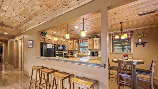 A kitchen with wooden cabinets, a central island with stools, pendant lighting, and a dining area with chairs and a table. - Cabin in the Valley! Easy Access! Pet Friendly! (Sevierville)