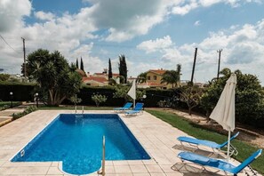 A rectangular swimming pool with a tiled deck, surrounded by blue lounge chairs and a white umbrella.