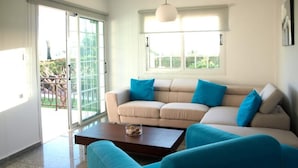 A living room with a beige sofa, blue cushions, a wooden coffee table, and a large window with a view of greenery. - Luxury, Modern Villas with private pool & garden in Coral Bay area (Πέγεια, Πάφος)