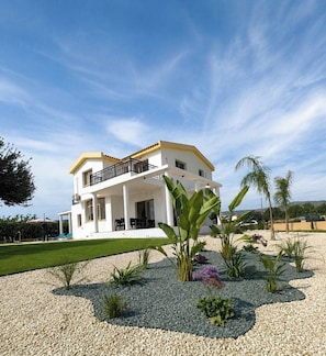 A two-story house with a balcony, surrounded by a landscaped garden with various plants and a gravel driveway. - Luxury, Modern Villa with private pool & garden in Coral Bay area (Πέγεια, Πάφος)