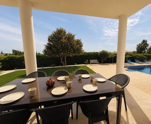 An outdoor dining area with a table set for six, surrounded by chairs, with a pool and greenery in the background. - Luxury, Modern Villa with private pool & garden in Coral Bay area (Πέγεια, Πάφος)