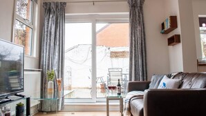A living room with a sofa, a glass coffee table, and a TV on a stand. There is a sliding glass door leading to an outdoor area with a bicycle and a table. - Bedford Road Garden Apartment (Bedford)