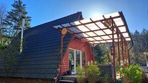 Una cabaña de madera con techo inclinado, un porche con pérgola y un pequeño jardín con plantas en macetas y una manguera.