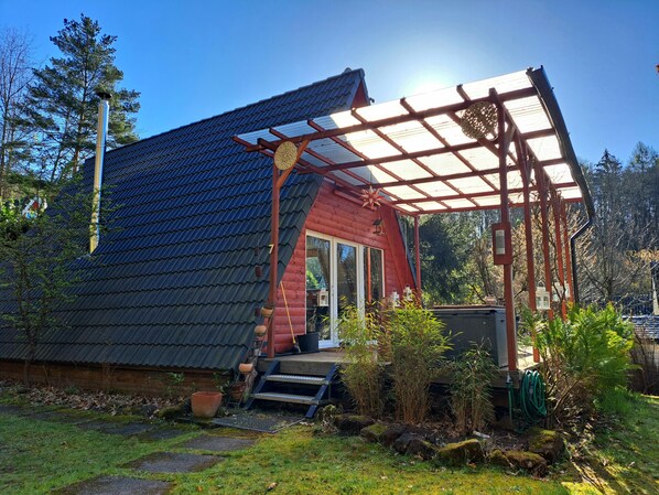 A wooden cabin with a sloped roof, a porch with a pergola, and a small garden area with potted plants and a hose.