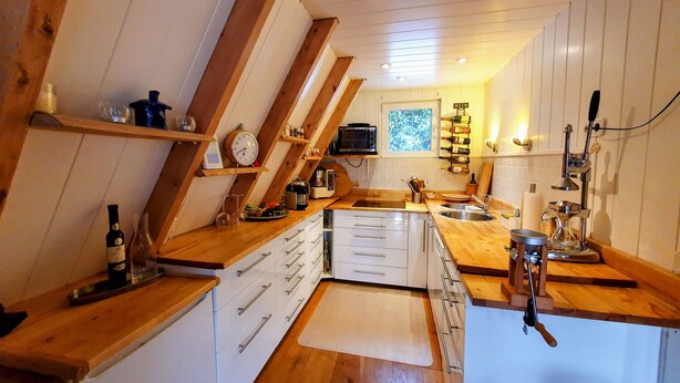 Cocina compacta con mostradores de madera, armarios blancos y estufa integrada. Hay botellas de vino y copas en los estantes, y un reloj en la pared.