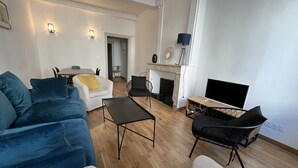 Een moderne zithoek met een blauwe bank, een witte fauteuil, een zwarte salontafel en een televisie op een houten dressoir.