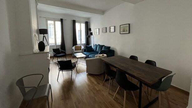 Een ruime woonkamer met een houten eettafel, een bank en een stoel. Er hangen afbeeldingen in lijsten aan de muur en een raam met gordijnen.