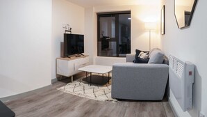 A modern living room with a grey sofa, a white coffee table, a flat-screen TV, a small side table, a floor lamp, and a large window. - Contemporary 1 Bed Apartment in Central Liverpool (Liverpool)