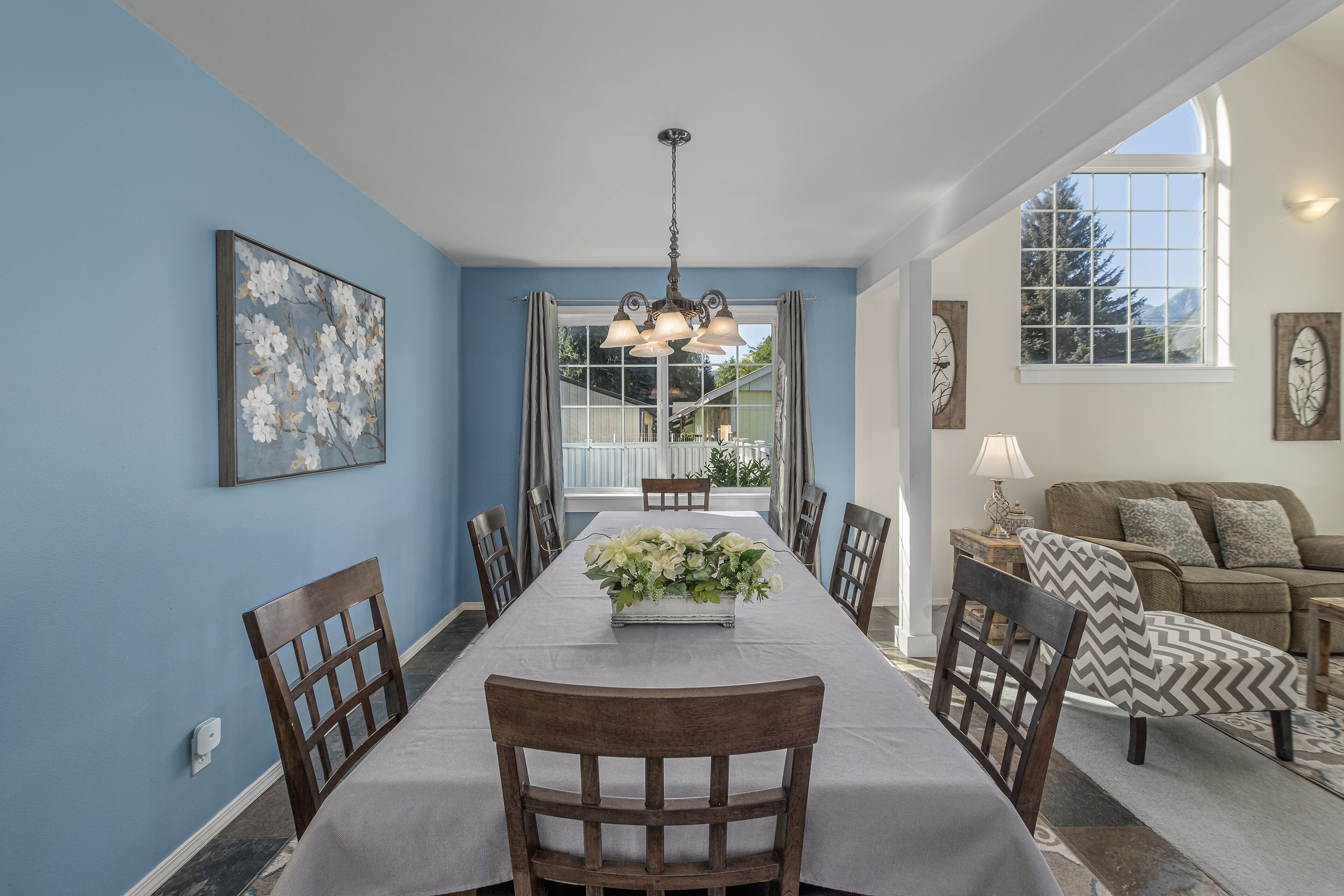 Une salle à manger avec une longue table, des chaises, un lustre et une fenêtre donnant sur des arbres et des montagnes.