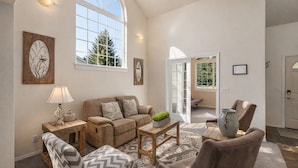 Ein gemütliches Wohnzimmer mit Sofa, Sesseln, Couchtisch und Lampe. Zwei Fenster und eine Tür führen auf einen Balkon.