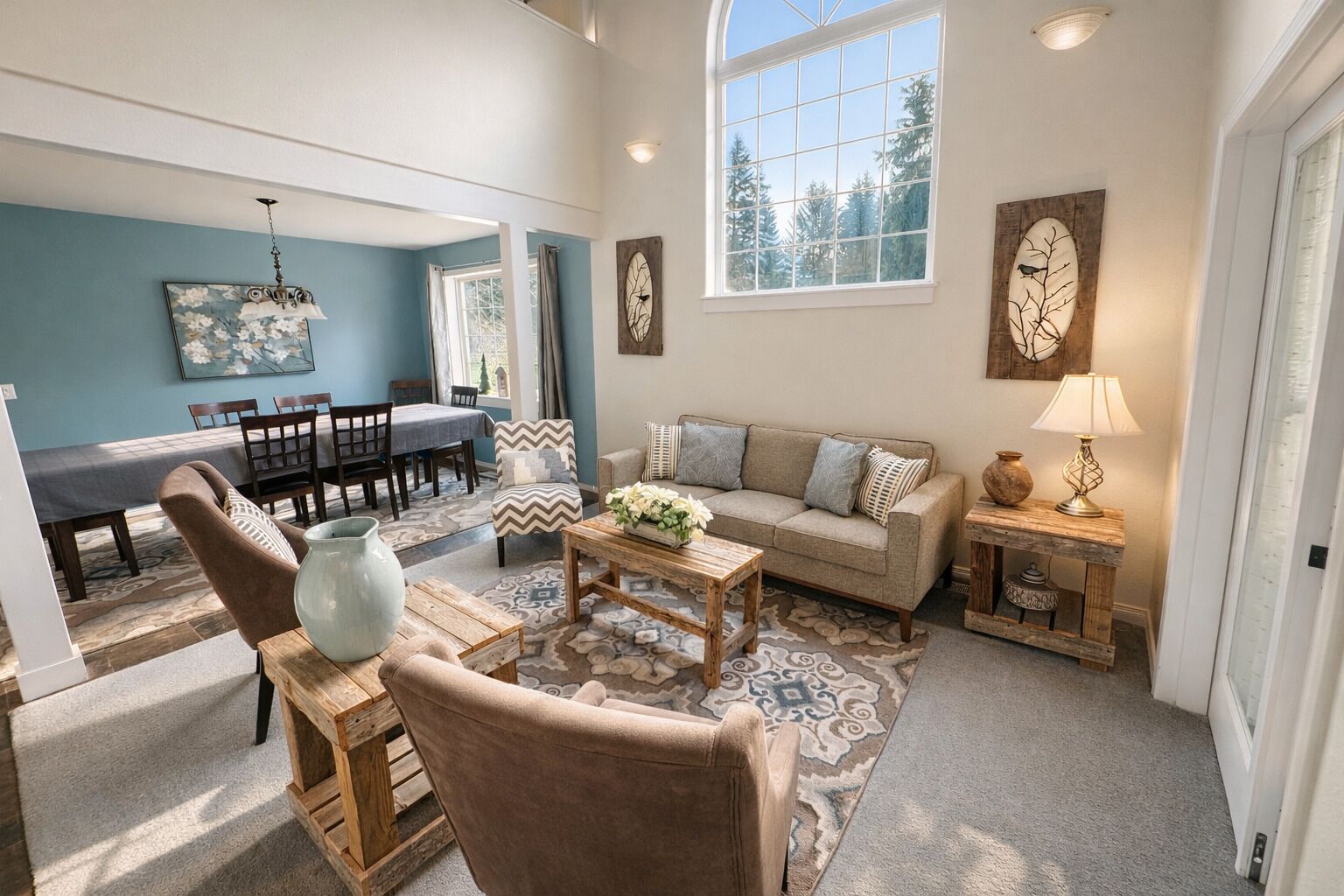 A spacious living room with a sofa, coffee table, and decorative items. There is a dining area with a table and chairs, and a large window providing natural light.