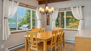A dining area with a wooden table and chairs, large windows with curtains, a chandelier, and a view of a lake and forest.