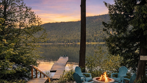 A lakeside deck with a fire pit and Adirondack chairs overlooking a forested lake.