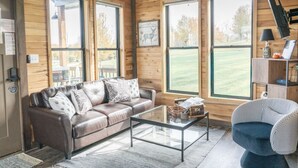 A cozy cabin interior with a leather sofa, a glass coffee table, and a houndstooth armchair.